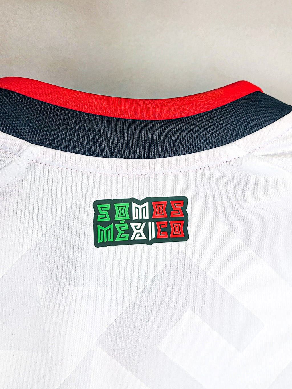 Close-up of a white sports jersey with a red collar and 'Somos Mexico' logo on a light gray background.